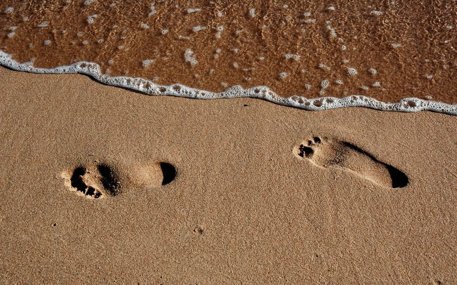 Footprints in wet sand beside a small breaking wave.